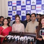 A group of people, including a woman in a white lace dress and a woman in a red saree, standing in front of a backdrop with various logos and brands, giving a press conference with multiple microphones on a podium.