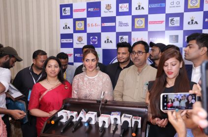 A group of people, including a woman in a white lace dress and a woman in a red saree, standing in front of a backdrop with various logos and brands, giving a press conference with multiple microphones on a podium.